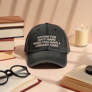 Peut inclure: Casquette de baseball gris foncé avec le texte blanc « HAVING FUN ISN'T HARD WHEN YOU HAVE A LIBRARY CARD ». Entourée de livres, de lunettes et d'une bougie allumée, suggérant le thème de la lecture.