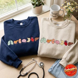 Può includere: Una felpa blu navy e una felpa beige, ciascuna con una fila ricamata di organi umani in stile cartoon che si tengono per mano, inclusi polmoni, pancreas, reni, cervello, stomaco, cuore e fegato. Uno stetoscopio è posizionato nelle vicinanze.