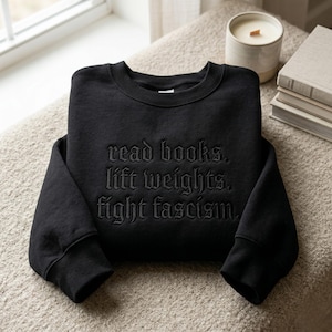 Könnte beinhalten: Schwarzes Sweatshirt mit Rundhalsausschnitt und dem gestickten Text: "read books. lift weights. fight fascism." Das Sweatshirt liegt gefaltet auf einer neutralfarbenen Oberfläche, mit einer Kerze und Büchern im Hintergrund.