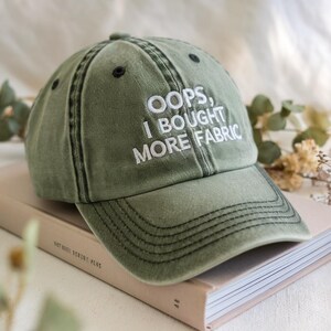 Peut inclure: Casquette de baseball vert olive avec le texte brodé "OOPS, I BOUGHT MORE FABRIC". La casquette a une visière incurvée, des coutures noires et un aspect vieilli. Le chapeau est posé sur un livre.