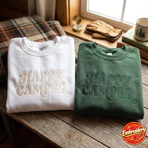 Könnte beinhalten: Ein weißes Sweatshirt und ein dunkelgrünes Sweatshirt, beide mit dem gestickten Text "HAPPY CAMPER" in einer wellenförmigen Retro-Schriftart. Das weiße Sweatshirt hat eine hellbeige Stickerei, während das grüne eine dunkelgrüne Stickerei aufweist.
