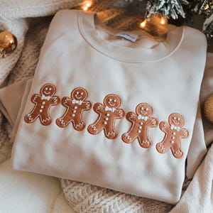 Może przedstawiać: Kremowa bluza z haftowanym wzorem pięciu piernikowych ludzików. Każdy piernikowy ludzik ma białą muszkę, uśmiechniętą twarz i brązowe detale. Bluza jest złożona na teksturowanej powierzchni.