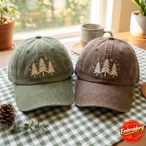 Può includere: Due cappellini da baseball, uno verde oliva e uno marrone, ciascuno con un ricamo raffigurante tre alberi di pino circondati da piccole stelle. I cappellini presentano una corona strutturata, una visiera curva e occhielli cuciti.