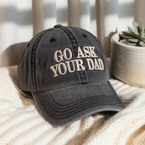 Puede incluir: Una gorra de béisbol de algodón negro lavado con el texto "Go Ask Your Dad" bordado en blanco.