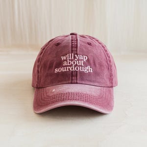 May include: A maroon baseball cap with the phrase "will yap about sourdough" embroidered in white. The cap has a curved brim and an adjustable strap at the back. The hat is made of a soft, worn material.