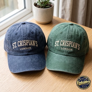 Puede incluir: Dos gorras de béisbol, una azul marino y otra verde bosque, ambas con un acabado de tela desgastada. Cada gorra tiene el texto bordado ST. CRISPIAN'S LONDON en un color crema claro en el panel frontal sobre la visera curva.
