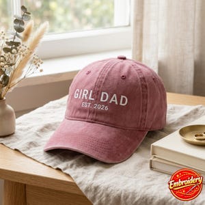 May include: A dusty rose baseball cap featuring white embroidered text that reads GIRL DAD EST. 2026. The cap has a structured crown, a curved brim, and stitched eyelets on the top panels, showcasing a washed fabric texture throughout the design.