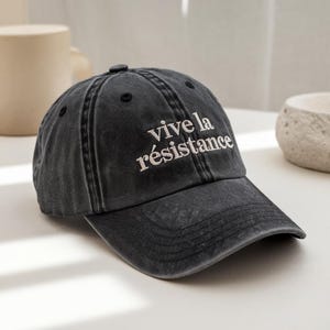 May include: A dark grey baseball cap with the embroidered text "Vive la résistance" in white. The cap has a curved peak and a distressed, vintage appearance. The hat is made of a soft, durable fabric.
