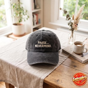 May include: A charcoal gray baseball cap with a curved brim and an embroidered design on the front. The text on the cap reads "PAUSE... NEVERMIND." in a light-colored, sans-serif font. The cap features a distressed, washed-out fabric texture.