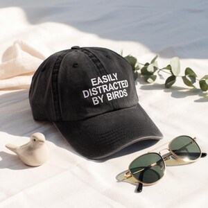 May include: A black baseball cap with the text "EASILY DISTRACTED BY BIRDS" in white. The image also includes gold-rimmed sunglasses and a small ceramic bird figurine.