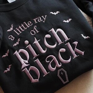 May include: Black sweatshirt featuring the embroidered phrase "a little ray of pitch black", bats, and a coffin. The text and bats are in a light purple colour. The sweatshirt is a crew neck style.