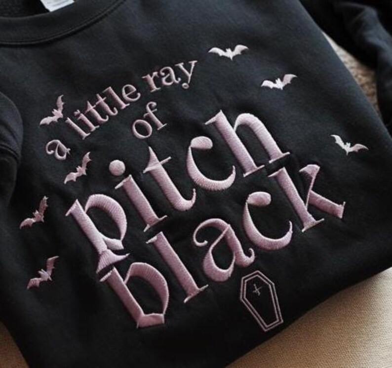 May include: Black sweatshirt featuring the embroidered phrase "a little ray of pitch black", bats, and a coffin. The text and bats are in a light purple colour. The sweatshirt is a crew neck style.