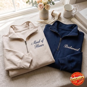 May include: A beige quarter-zip sweatshirt with the embroidered text Maid of Honor and a navy blue quarter-zip sweatshirt with the embroidered text Bridesmaid. Both garments feature a high collar, a metal zip, and ribbed sleeve cuffs.