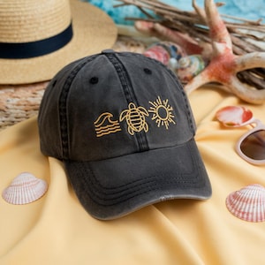 Pode incluir: Boné de beisebol preto desgastado com desenhos amarelos bordados de ondas, uma tartaruga e um sol. O boné está sobre uma superfície amarela com um chapéu de palha, conchas e óculos de sol no fundo.