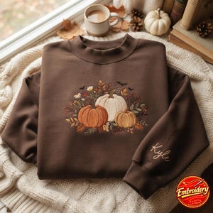 May include: A dark brown crewneck sweatshirt featuring an embroidered design of three pumpkins in orange, white, and tan, surrounded by autumn leaves, berries, and small bats. The name Kyle is embroidered in cursive on the sleeve cuff.