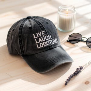 May include: A black baseball cap with white embroidered text that reads "LIVE LAUGH LOBOTOMY".