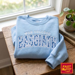 Puede incluir: Una sudadera azul claro con la inscripci&oacute;n bordada "WAR COLLEGE BASCIATH" en blanco y un dise&ntilde;o floral azul. La prenda est&aacute; doblada sobre una mesa de madera, cerca de una ventana y una peque&ntilde;a planta en maceta.