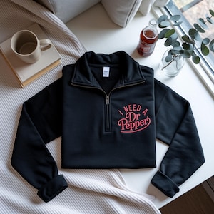 Puede incluir: Sudadera negra con cremallera de un cuarto con el texto "I NEED Dr Pepper" en escritura rosa. Una botella de refresco Dr Pepper es visible en el fondo, junto con una taza y un libro.