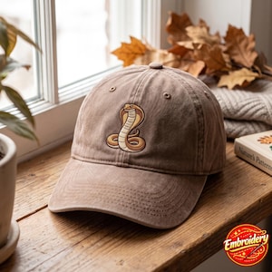 Puede incluir: Una gorra de béisbol marrón con un gráfico bordado de una cobra enroscada en el panel frontal. La gorra tiene una corona estructurada, una visera curva y ojales cosidos. El diseño muestra una cobra de color beige y marrón.