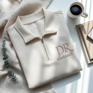 Könnte beinhalten: Cremefarbenes Sweatshirt mit Kragen und einem Viertel-Reißverschluss. Der gestickte Text "DR darnell" ist auf der Vorderseite zu sehen. Eine Tasse Kaffee und ein Stapel Bücher sind im Hintergrund.