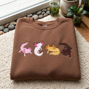 May include: A brown crewneck sweatshirt featuring an embroidered design of four axolotls in a row. The axolotls are depicted in distinct colours: light pink, white with pink gills, golden yellow, and dark brown, each with detailed stitching.