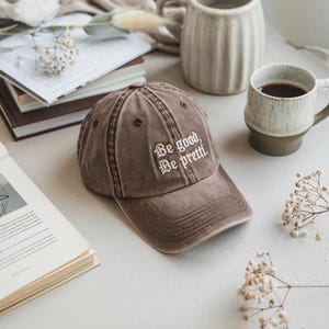 May include: A brown baseball cap with the embroidered text "Be good, Be pretti." The cap is on a white surface with books, mugs, and decorative flowers.