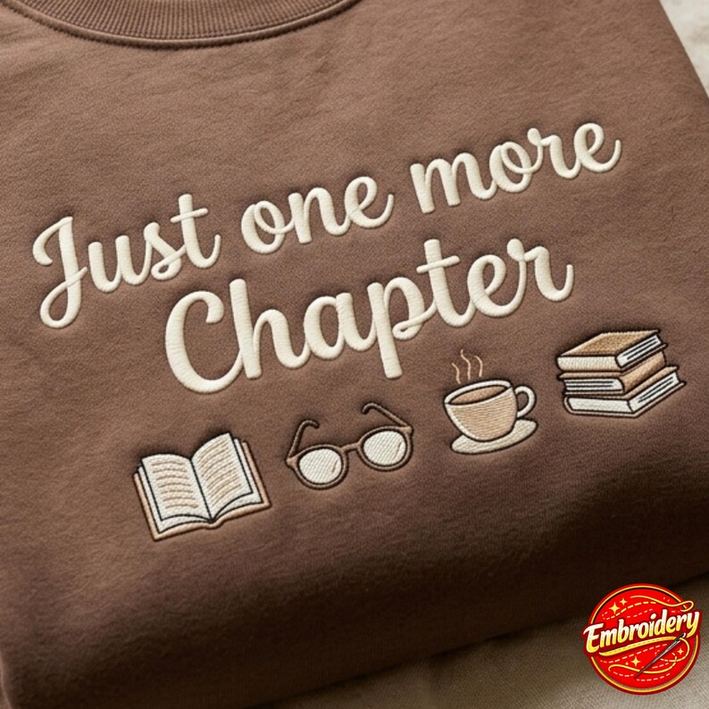 May include: A brown sweatshirt featuring cream-colored embroidered text that reads "Just one more Chapter" above a series of embroidered icons including an open book, a pair of round glasses, a steaming cup of coffee, and a small stack of books.