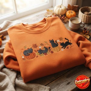 Pode incluir: Uma camisola laranja de gola redonda com um design bordado no peito. O bordado retrata quatro gatos pretos a interagir com abóboras cor de laranja, rodeados por várias folhas de outono e pequenas flores brancas em tons de dourado e castanho.