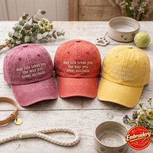 Könnte beinhalten: Drei Baseballkappen in Altrosa, Terrakotta und Senfgelb. Jede Kappe ist mit dem weißen Sticktext versehen: may life treat you the way you treat animals. Die Kappen haben eine strukturierte Krone, einen gebogenen Schirm und einen Knopf oben.