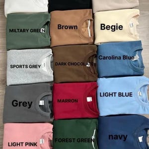 Puede incluir: Una colecci&oacute;n de sudaderas de cuello redondo dobladas en varios colores, incluyendo blanco, negro, arena, verde militar, marr&oacute;n, beige, gris deportivo, chocolate oscuro, azul Carolina, gris, granate, azul claro, rosa claro, verde bosque y marino. Cada camisa tiene una etiqueta.