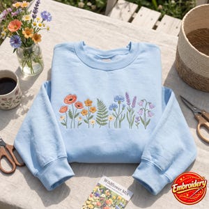 May include: A light blue crewneck sweatshirt featuring a central embroidered design of various wildflowers including orange poppies, yellow daisies, blue cornflowers, green ferns, and purple lavender sprigs, all rendered in detailed thread work.