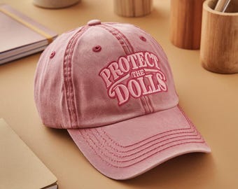 Puede incluir: Una gorra de béisbol rosa con el texto "Protect the Dolls" bordado en rosa en la parte delantera.