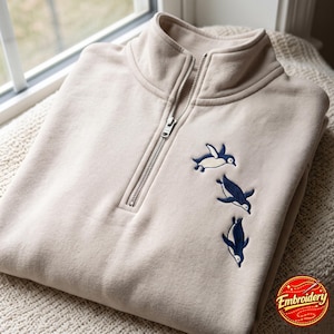 Könnte beinhalten: Ein beigefarbenes Sweatshirt mit kurzem Reißverschluss und einer Stickerei von drei Pinguinen in Marineblau und Weiß. Das Kleidungsstück verfügt über einen Stehkragen mit einem silberfarbenen Metallreißverschluss an der Brust.