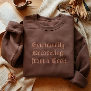Könnte beinhalten: Ein braunes Sweatshirt mit dem gestickten Text "Emotionally Recovering from a Book". Das Sweatshirt wird auf einer neutralfarbenen Decke präsentiert, mit einem Buch und einer Tasse Kaffee im Hintergrund. Der Text ist in einer dekorativen Schriftart.
