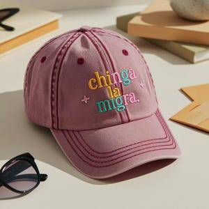 Può includere: Un cappellino da baseball rosa polveroso con la scritta ricamata "Chinga la migra" in giallo, verde acqua e rosa. Il cappellino ha una visiera curva, dettagli cuciti e piccoli accenti marroni. Un paio di occhiali da sole si trova nell'angolo in basso a sinistra.