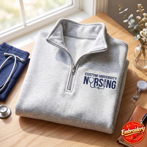 May include: A light gray quarter-zip pullover featuring dark blue embroidery on the chest that reads CUSTOM UNIVERSITY NURSING, with a stethoscope replacing the letter U and a syringe replacing the letter I in the word NURSING.