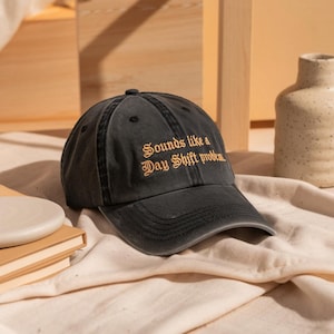 Könnte beinhalten: Eine schwarze Baseballkappe mit dem goldfarben gestickten Text "Sounds like a Day Shift problem". Die Kappe hat einen gebogenen Schirm und einen Used-Look. Der Hut liegt auf einem cremefarbenen Stoff.