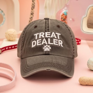 May include: A distressed brown baseball cap with the text "TREAT DEALER" in white, along with a white paw print. The cap has a curved brim and a classic six-panel design, perfect for dog lovers.
