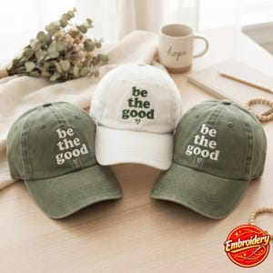 Könnte beinhalten: Drei Baseballkappen mit dem gestickten Text be the good und einem kleinen Herzsymbol. Eine Kappe ist weiß mit dunkelgrüner Schrift, während die anderen beiden olivgrün mit weißer Schrift sind. Jede Kappe hat einen gebogenen Schirm.
