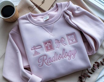 Puede incluir: Sudadera rosa claro con la palabra "Radiology" bordada en cursiva. La sudadera presenta imágenes bordadas de símbolos médicos. Una taza de café y un collar son visibles en el fondo.