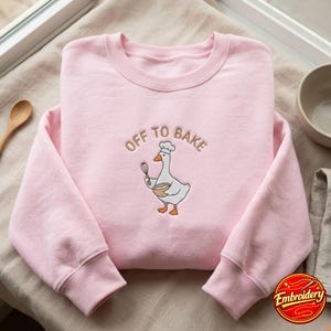 May include: A light pink crewneck sweatshirt featuring an embroidered design of a white goose wearing a chef hat, holding a whisk and a mixing bowl. The text "OFF TO BAKE" is embroidered in a brown arc above the goose illustration.