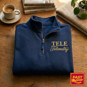 May include: A navy blue pullover sweatshirt with a quarter-zip collar and gold embroidered text that reads "TELE Telemetry." The sweatshirt is folded on a wooden surface, with a coffee cup and books in the background.