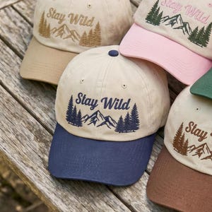 May include: Baseball caps in various colors, including tan, pink, and green, are displayed. Each cap features the embroidered phrase "Stay Wild" above a mountain and tree design. The caps have curved brims and adjustable straps.