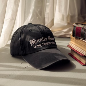 Puede incluir: Gorra de béisbol gris oscuro con la frase "Morally Grey is my favorite color" bordada en blanco. La gorra tiene una visera curva y un botón en la parte superior. Está sobre una superficie gris claro.