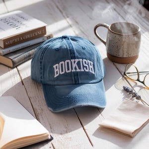 Pode incluir: Boné de beisebol azul denim com a palavra "BOOKISH" bordada em branco. O boné está sobre uma superfície de madeira branca, junto a livros, uma caneca e óculos. O boné tem um aspeto desgastado.