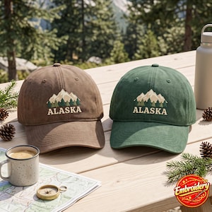 May include: Two baseball caps, one brown and one green, each featuring an embroidered design of beige mountains above dark green pine trees with the text ALASKA in beige lettering. The caps have a structured crown and a curved brim.