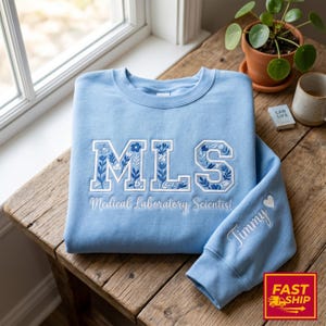 Op de afbeelding: Een lichtblauwe sweatshirt met de letters "MLS" geborduurd in wit met blauwe bloemmotieven. De woorden "Medical Laboratory Scientist" zijn eronder geborduurd. De mouw heeft de naam "Timmy" en een geborduurd hart.