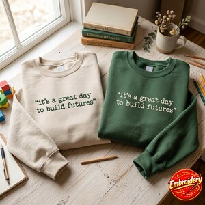May include: Two crewneck sweatshirts, one in beige and one in dark green, both featuring the embroidered text "it's a great day to build futures" in a dark font on the chest. The garments have long sleeves and ribbed cuffs and necklines.