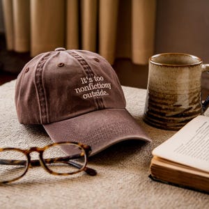 Puede incluir: Gorra de béisbol marrón con el texto bordado "it's too nonfictiony outside." La gorra está junto a una taza de cerámica, un par de gafas y un libro abierto. Los objetos están dispuestos sobre una superficie texturizada.