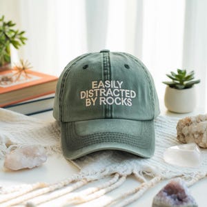 May include: A green baseball cap with the text "EASILY DISTRACTED BY ROCKS" embroidered in white on the front.